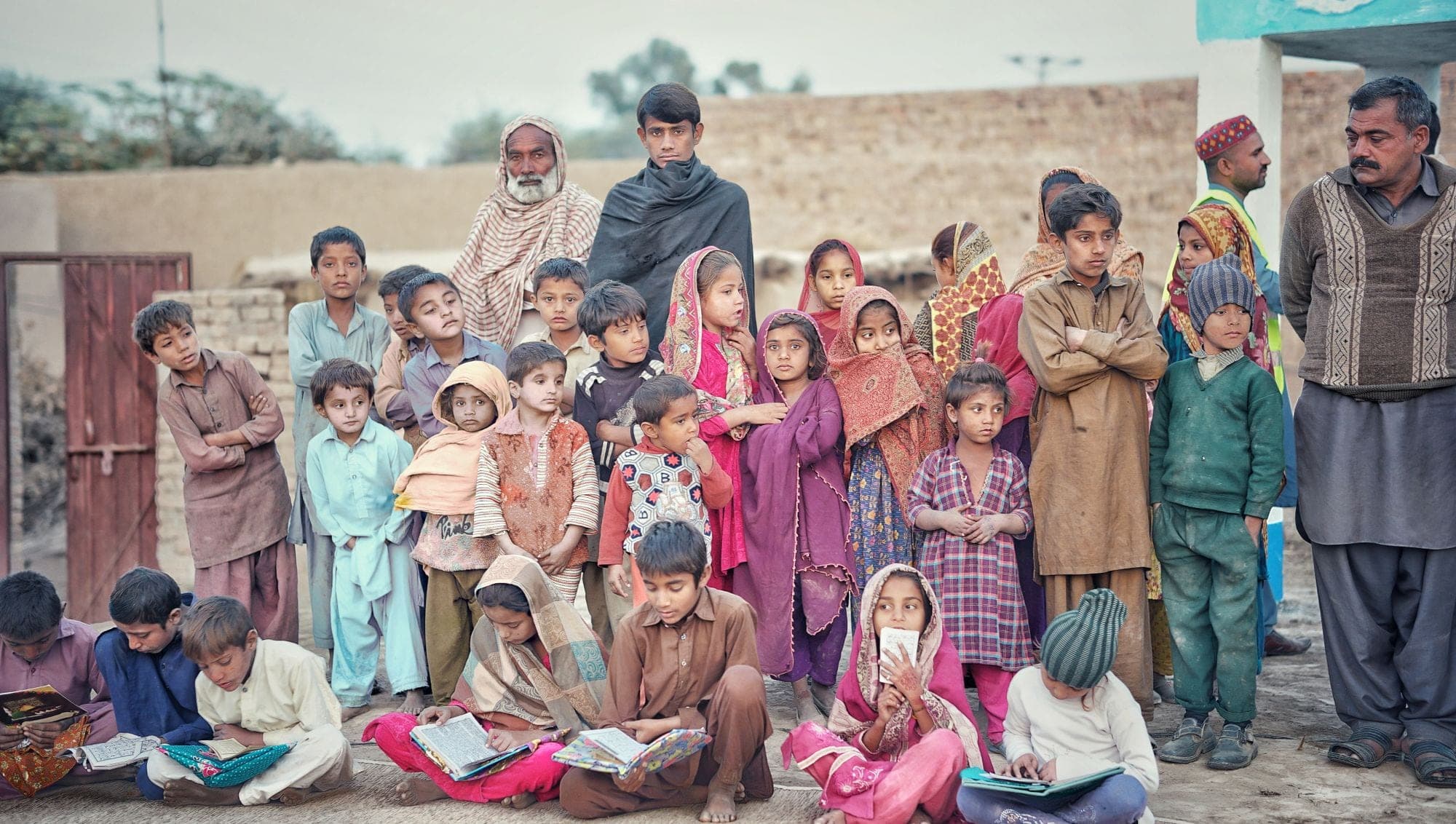 A large group of children and adults gathered for an outdoor class, some seated with books
